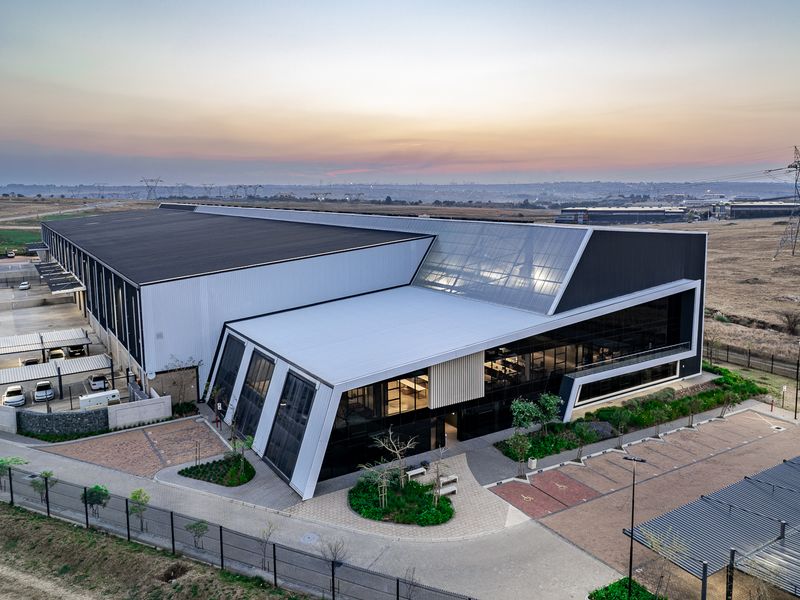 Aerial view of a modern industrial facility featuring a large warehouse structure, integrated office space with glass facade, and extensive paved parking areas.