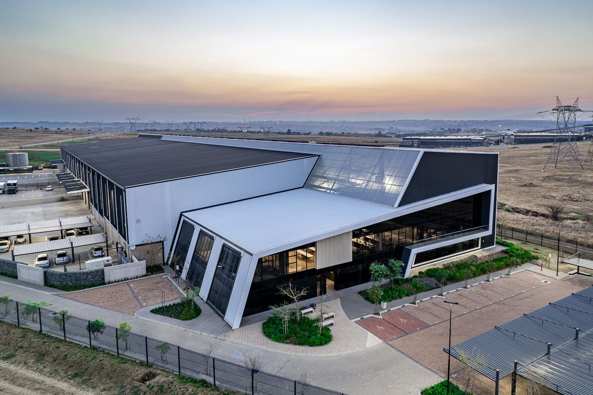 Aerial view of a modern industrial facility featuring a large warehouse structure, integrated office space with glass facade, and extensive paved parking areas.