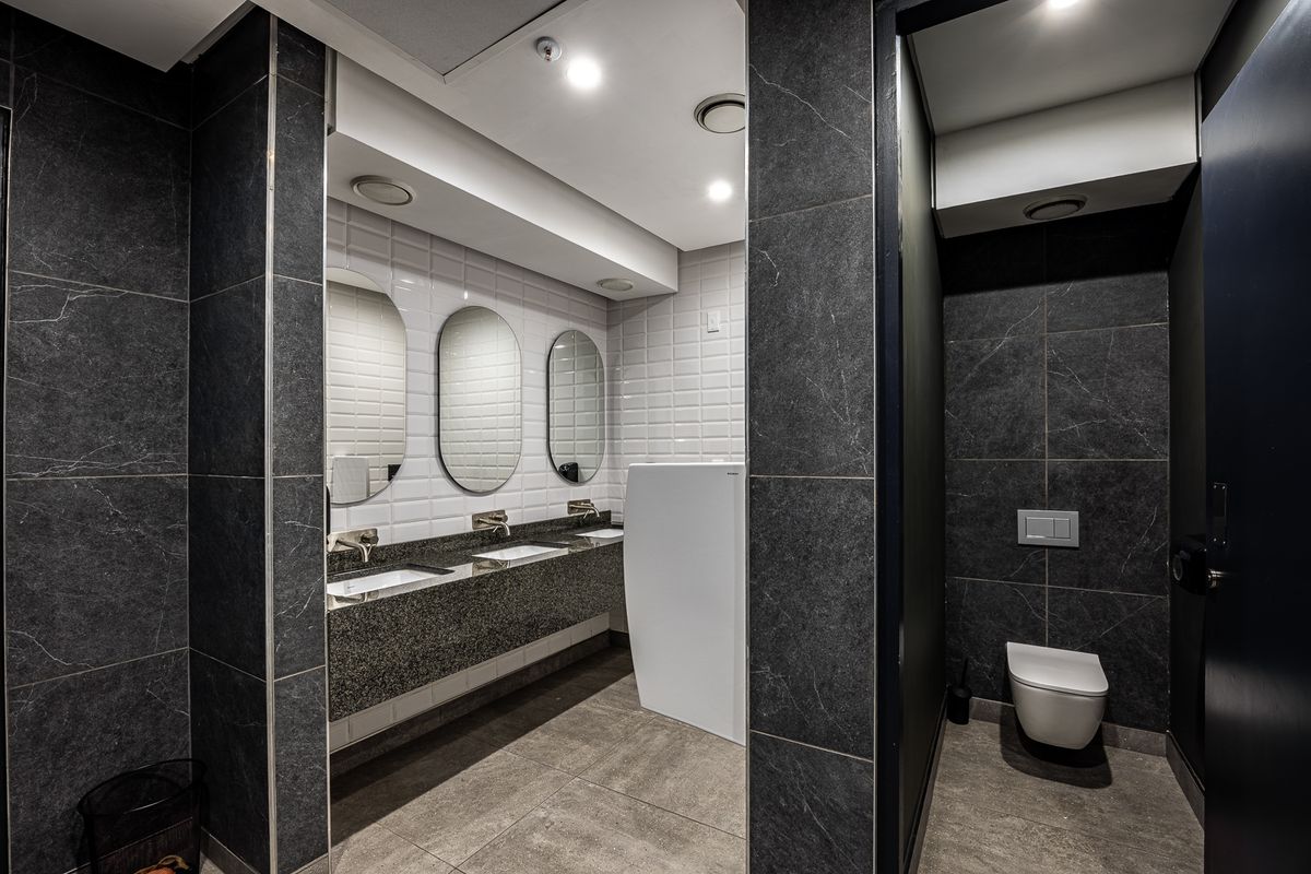 Modern industrial-style commercial bathroom featuring dark stone-look tiling, a triple vanity with integrated mirrors, and private wall-mounted toilet cubicles.