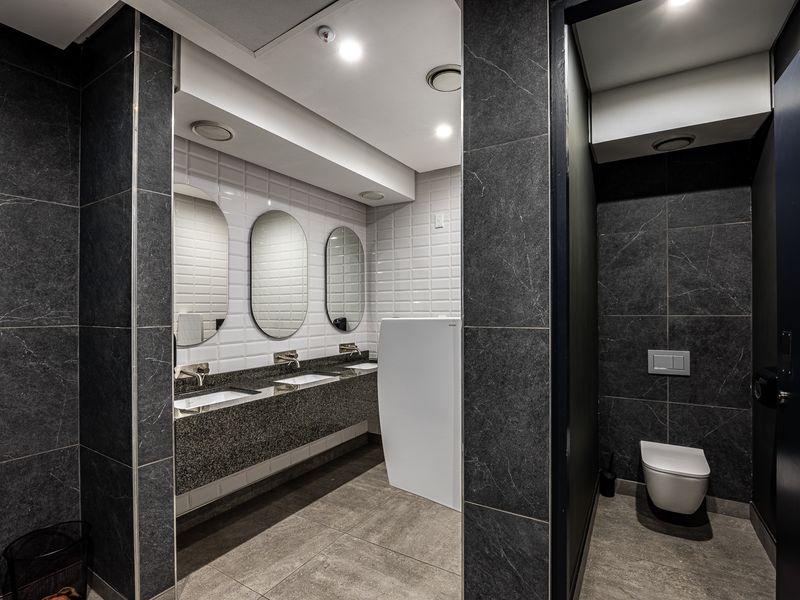 Modern industrial-style commercial bathroom featuring dark stone-look tiling, a triple vanity with integrated mirrors, and private wall-mounted toilet cubicles.