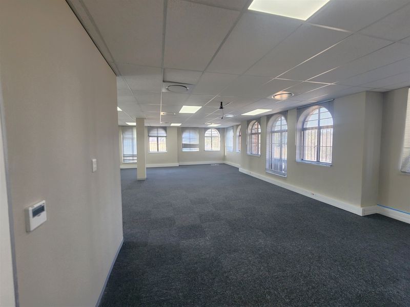 A spacious, vacant open-plan office area featuring carpeted flooring, a suspended ceiling with recessed lighting, and multiple arched windows providing natural light.