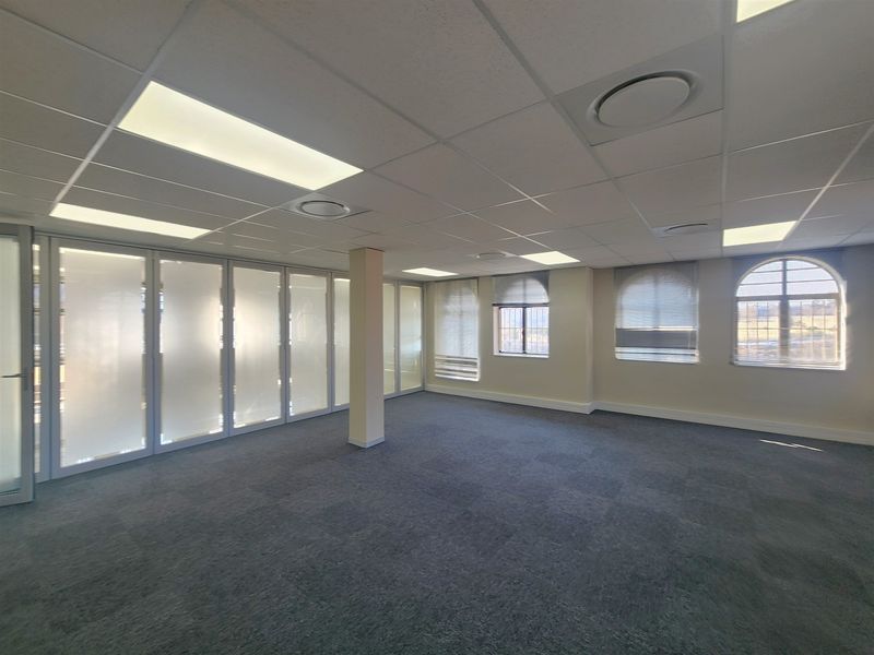 A spacious, vacant office area featuring a suspended ceiling, recessed lighting, glass partitions, and arched windows with integrated blinds.