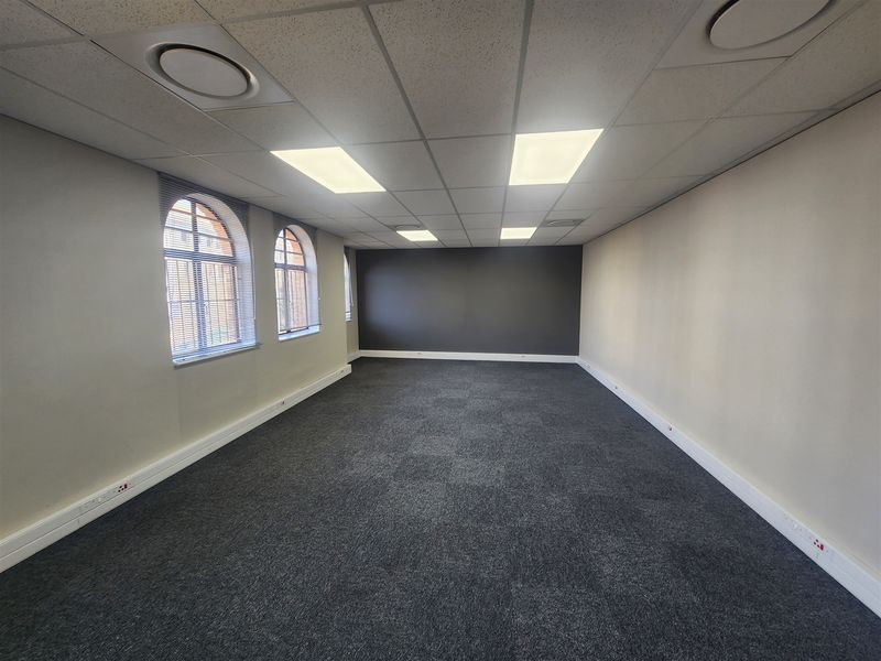 A clean, empty office space featuring carpet tiles, a suspended ceiling with recessed lighting, and arched windows with integrated power trunking.