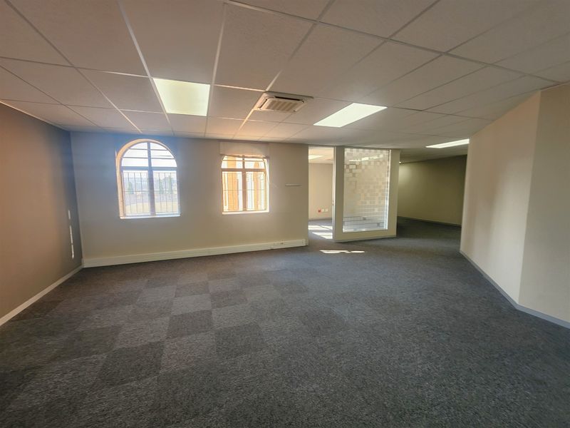 A spacious, empty office area featuring carpet tiles, a suspended ceiling with recessed lighting, ducted air conditioning, and arched windows with a glass partition.