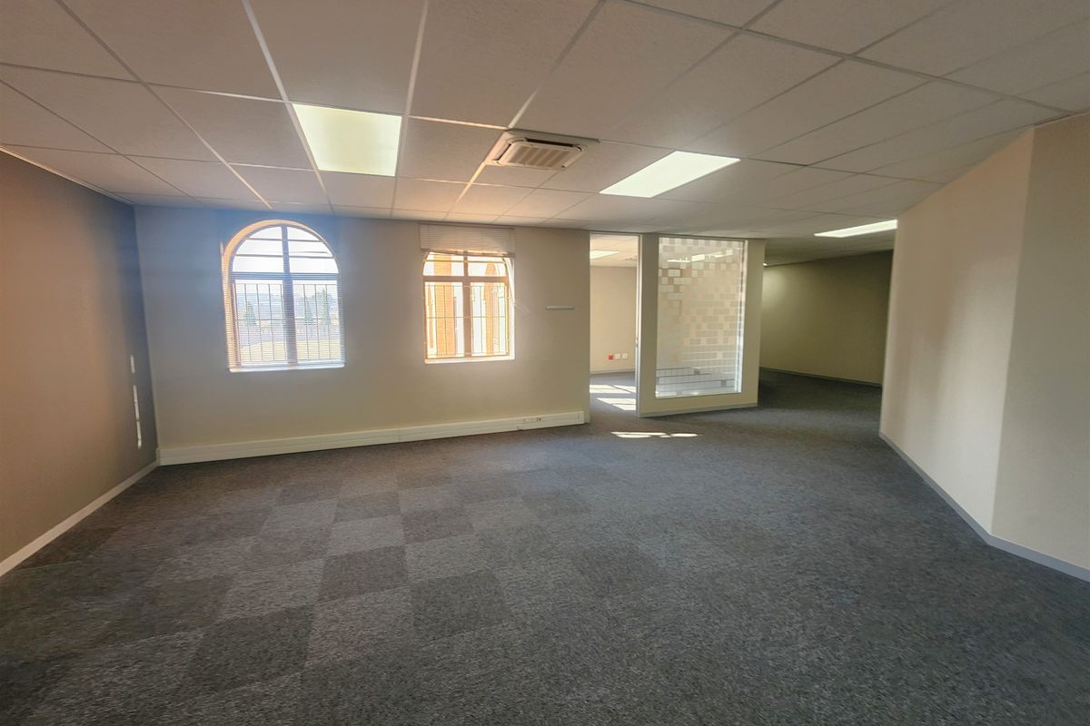 A spacious, empty office area featuring carpet tiles, a suspended ceiling with recessed lighting, ducted air conditioning, and arched windows with a glass partition.