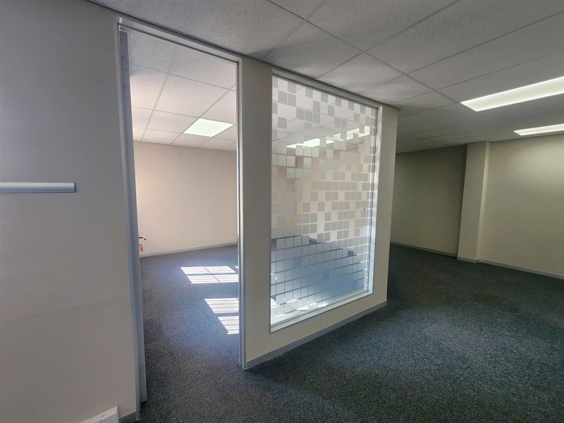 An empty office space featuring a glass partition with frosted privacy film, suspended ceiling tiles, recessed lighting, and neutral carpeted flooring.