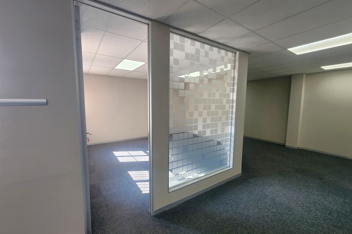 An empty office space featuring a glass partition with frosted privacy film, suspended ceiling tiles, recessed lighting, and neutral carpeted flooring.