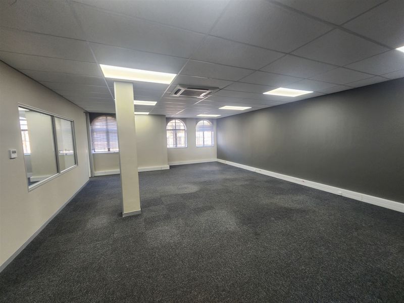 A spacious, empty open-plan office area featuring carpeted flooring, a suspended ceiling with recessed lighting, central air conditioning, and arched windows.