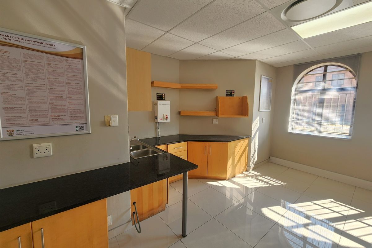 A clean, functional office kitchenette featuring granite countertops, wooden cabinetry, a stainless steel sink, and bright natural light from an arched window.