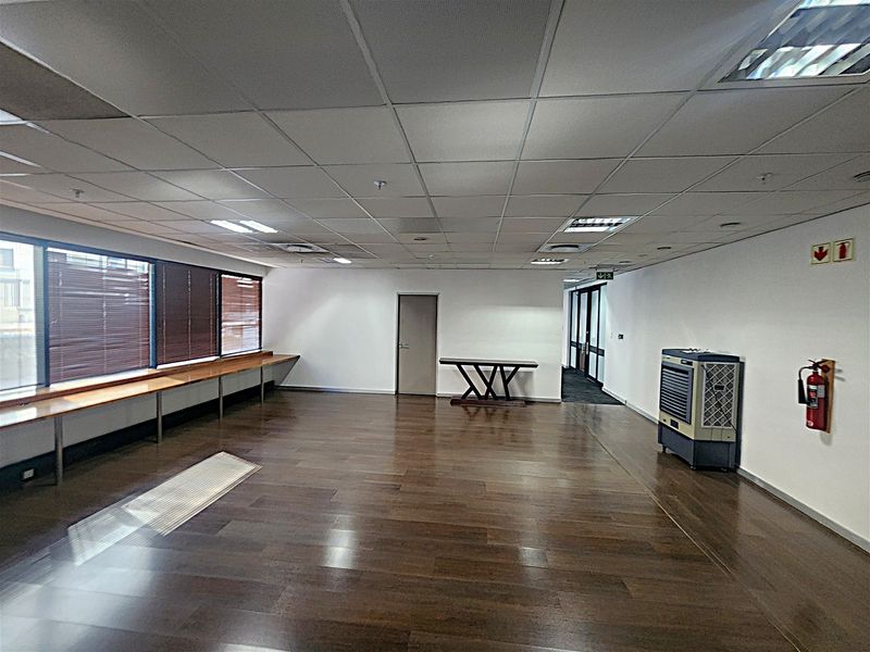 A spacious, open-plan office area featuring laminate flooring, a suspended ceiling with recessed lighting, and a long built-in desk along the window wall.
