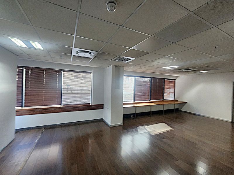An empty office space featuring hardwood flooring, a suspended ceiling with recessed lighting, and built-in desk surfaces along the windows.