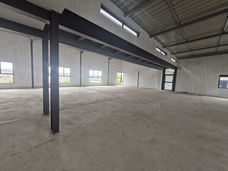 Spacious open-plan office area in shell condition, featuring exposed steel structural beams, concrete flooring, and large windows providing ample natural light.