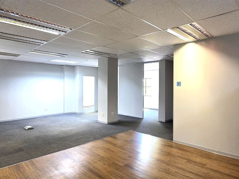 A spacious, empty office area featuring a mix of carpet and laminate flooring, suspended ceilings with recessed lighting, and a central structural column.