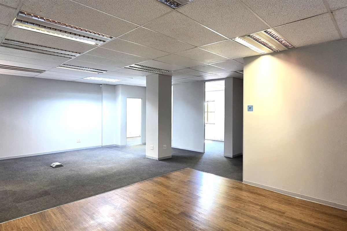 A spacious, empty office area featuring a mix of carpet and laminate flooring, suspended ceilings with recessed lighting, and a central structural column.