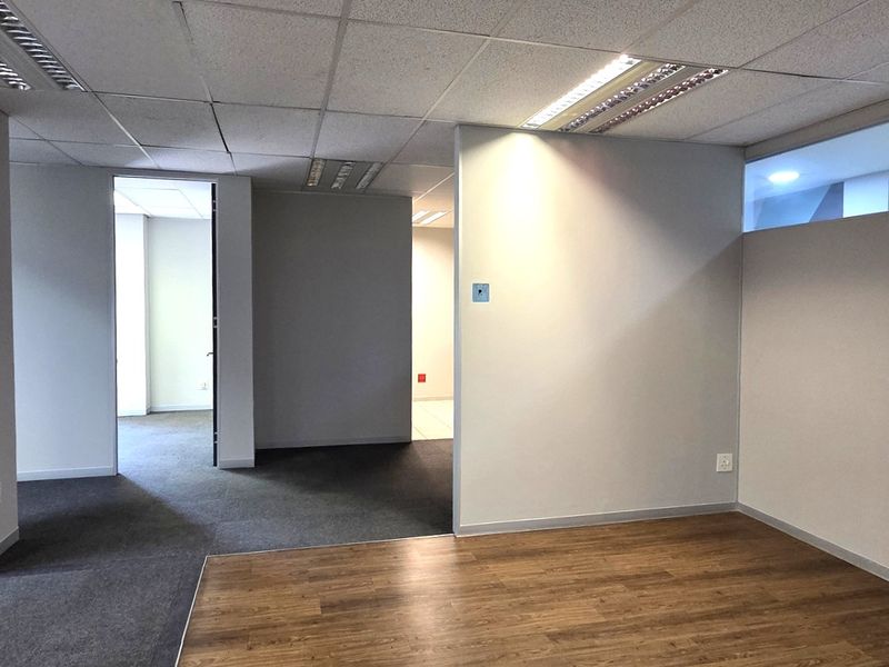 An empty, open-plan office space featuring a mix of laminate and carpet flooring, suspended ceilings with recessed lighting, and neutral partition walls.
