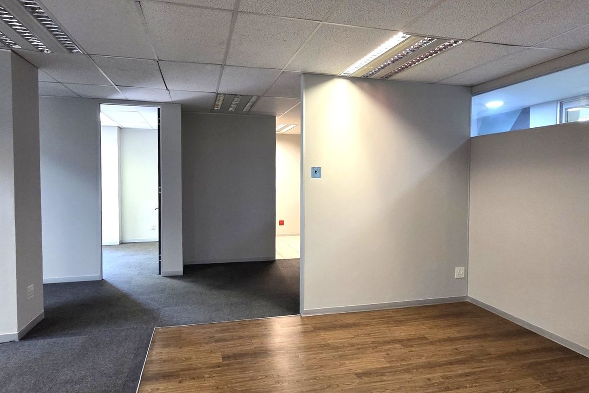 An empty, open-plan office space featuring a mix of laminate and carpet flooring, suspended ceilings with recessed lighting, and neutral partition walls.