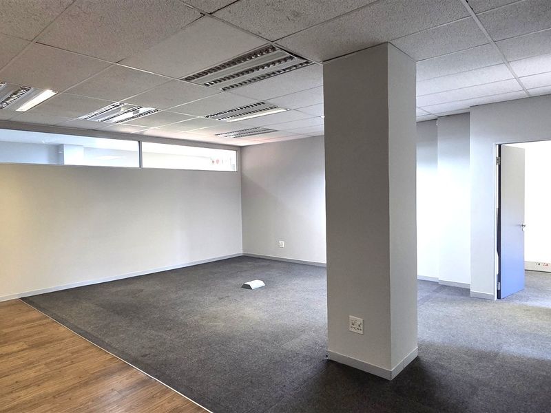 A spacious, vacant office area featuring a mix of carpet and laminate flooring, a suspended ceiling with recessed lighting, and a central structural column.