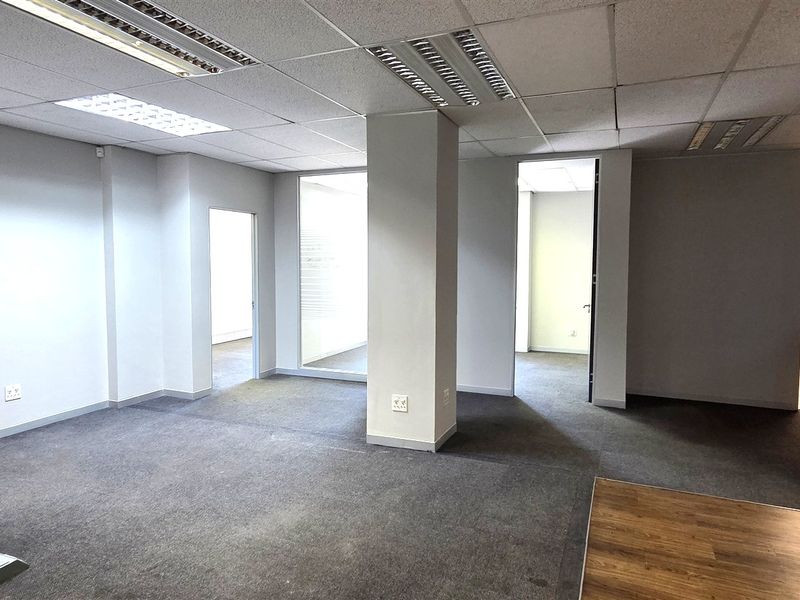A spacious, vacant office area featuring a mix of carpet and laminate flooring, suspended ceilings with recessed lighting, and a central structural column.