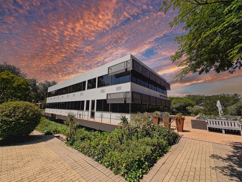 A modern two-story office building featuring tinted windows, external sun louvres, and a landscaped paved courtyard with a water feature.