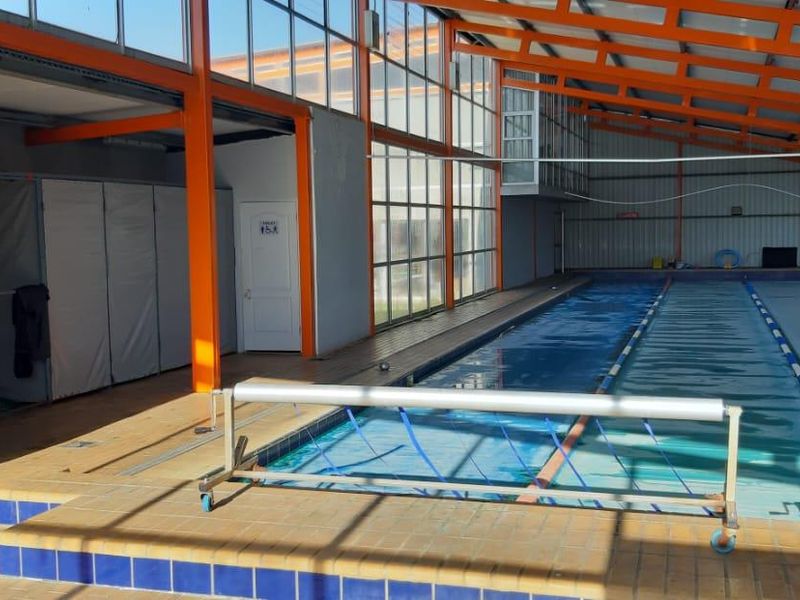 Indoor swimming pool facility featuring a multi-lane lap pool, large glass panel walls, and orange painted steel structural framing.