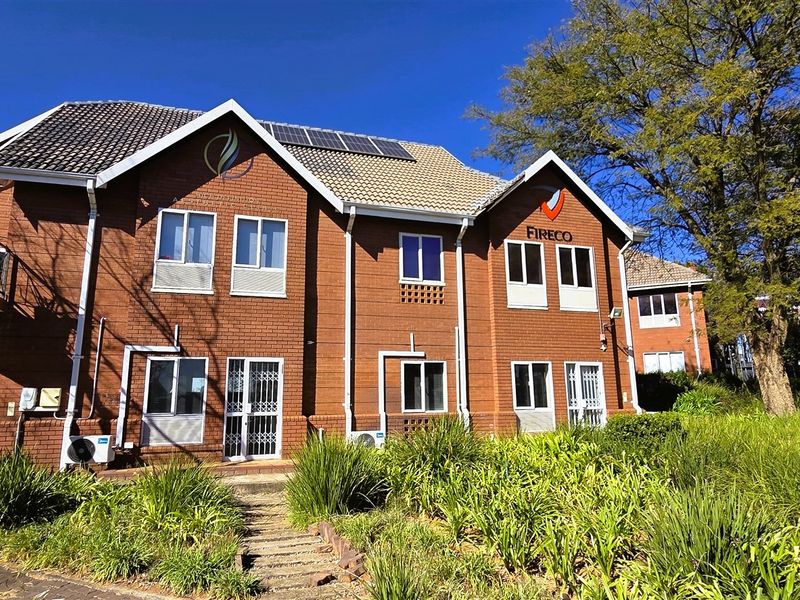 A well-maintained double-story brick office building featuring a tiled roof with solar panels, a balcony, and landscaped garden surroundings.