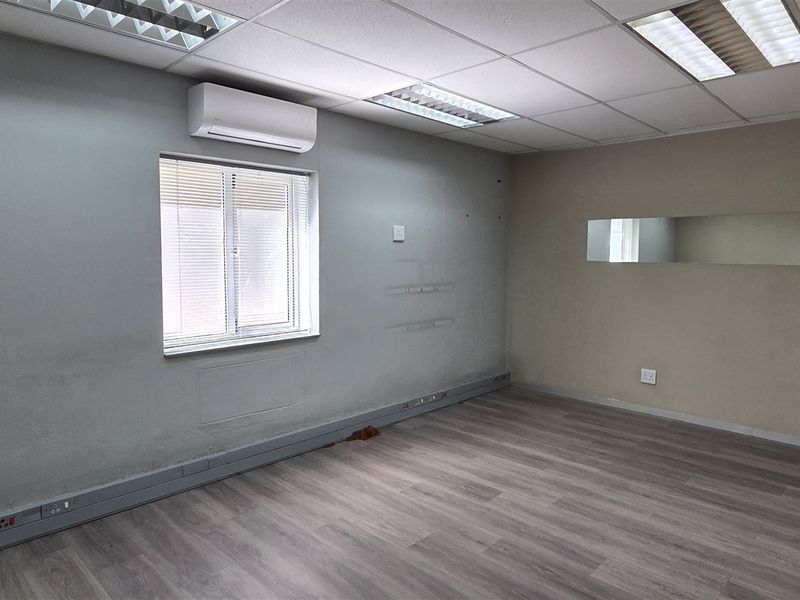 An empty office space featuring laminate flooring, a suspended ceiling with recessed lighting, and a wall-mounted air conditioning unit.