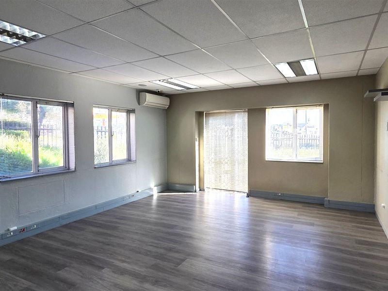 A spacious, empty office area featuring laminate flooring, a suspended ceiling with recessed lighting, and a wall-mounted air conditioning unit.
