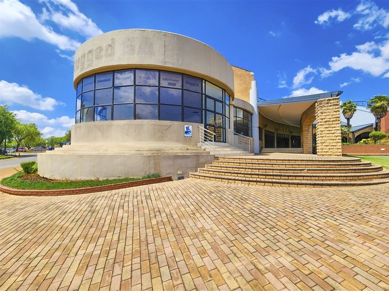 Modern office building exterior featuring a prominent curved glass facade, stone masonry accents, and extensive brick paving leading to the main entrance.