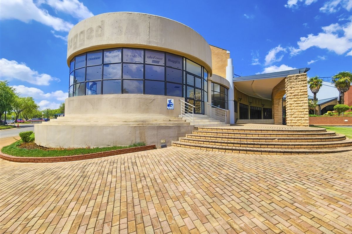 Modern office building exterior featuring a prominent curved glass facade, stone masonry accents, and extensive brick paving leading to the main entrance.