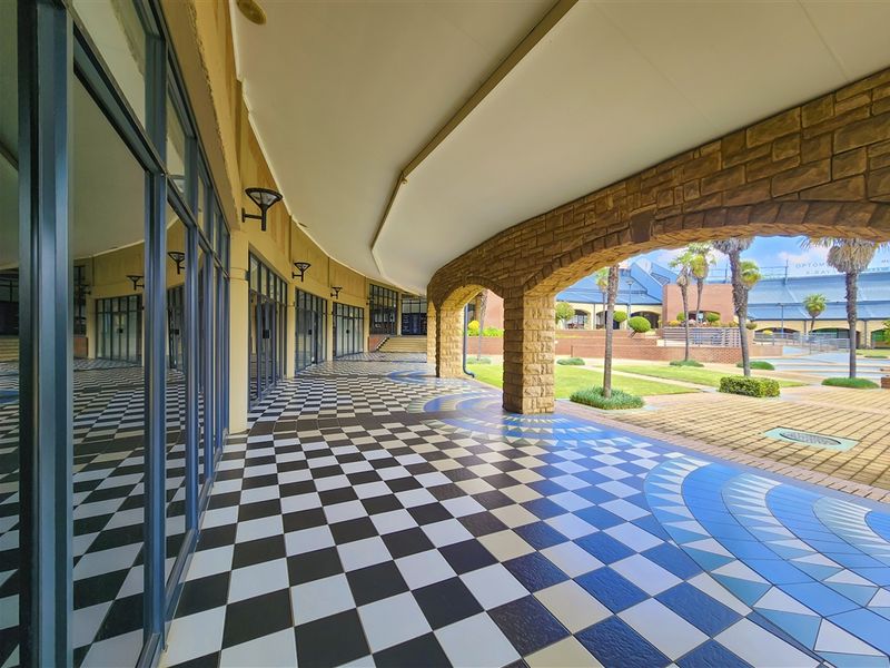 A spacious covered outdoor walkway featuring decorative checkered tile flooring, stone archways, and extensive glass frontage overlooking a landscaped courtyard.