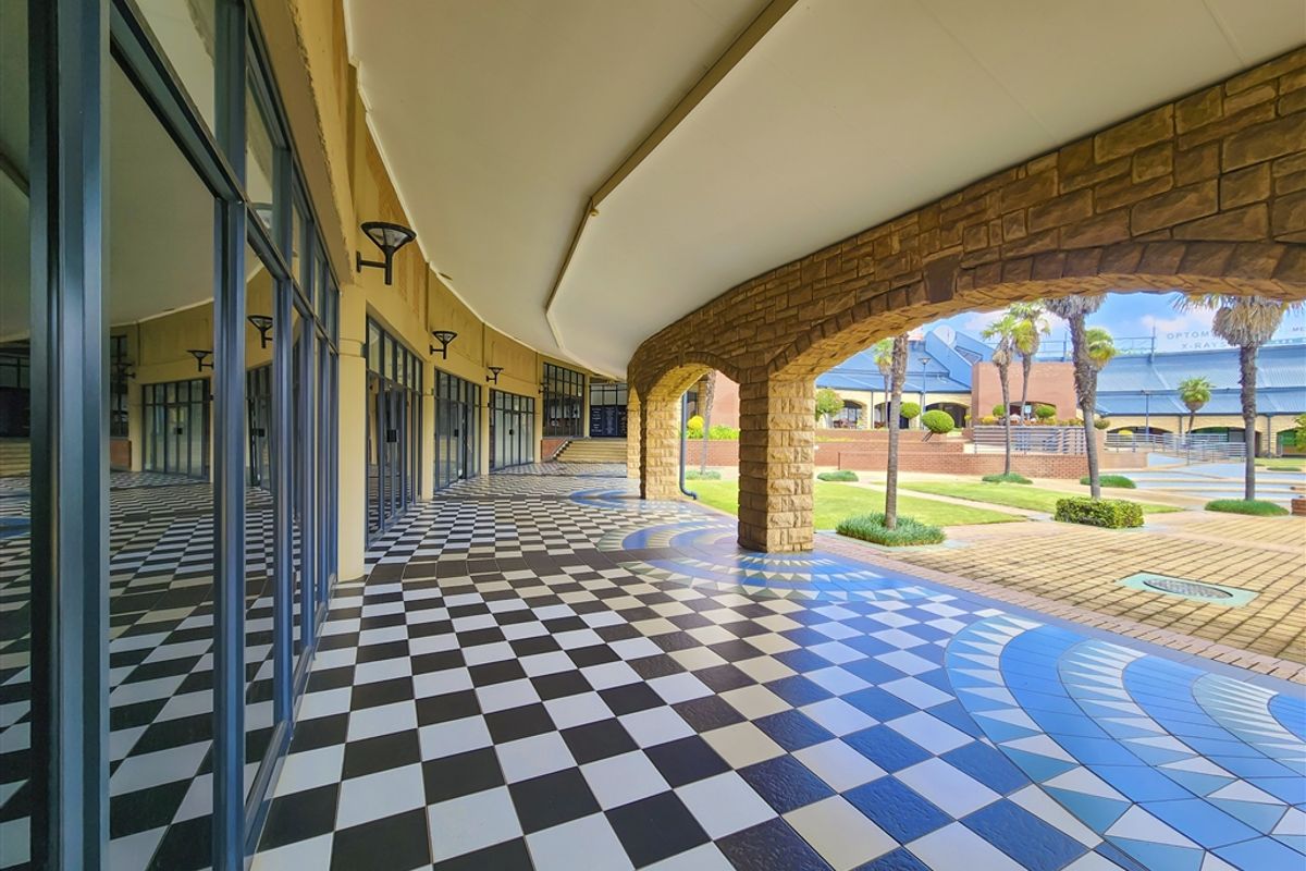 A spacious covered outdoor walkway featuring decorative checkered tile flooring, stone archways, and extensive glass frontage overlooking a landscaped courtyard.