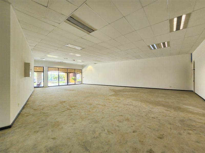 A spacious, empty open-plan office area featuring a suspended ceiling with recessed lighting and large windows providing natural light.