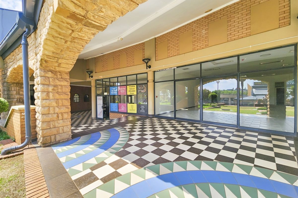A covered outdoor walkway featuring a decorative stone archway, patterned checkered tile flooring, and large glass storefront windows.
