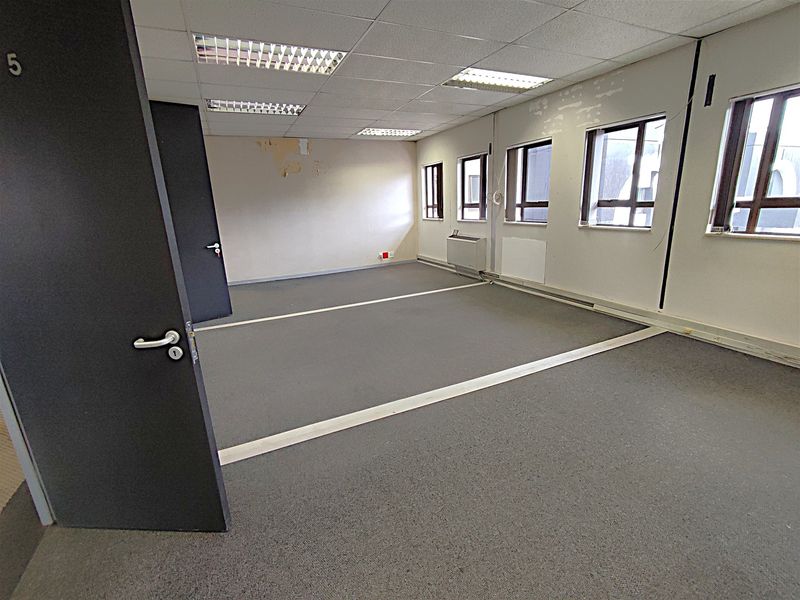 An empty office space featuring a suspended ceiling, recessed lighting, carpeted flooring, and wall-mounted HVAC unit, requiring cosmetic updates to the walls and flooring.