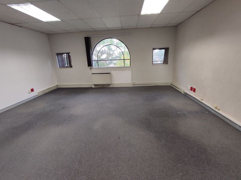 An empty office space featuring a suspended ceiling, recessed lighting, carpeted flooring, and an arched window with a wall-mounted HVAC unit.