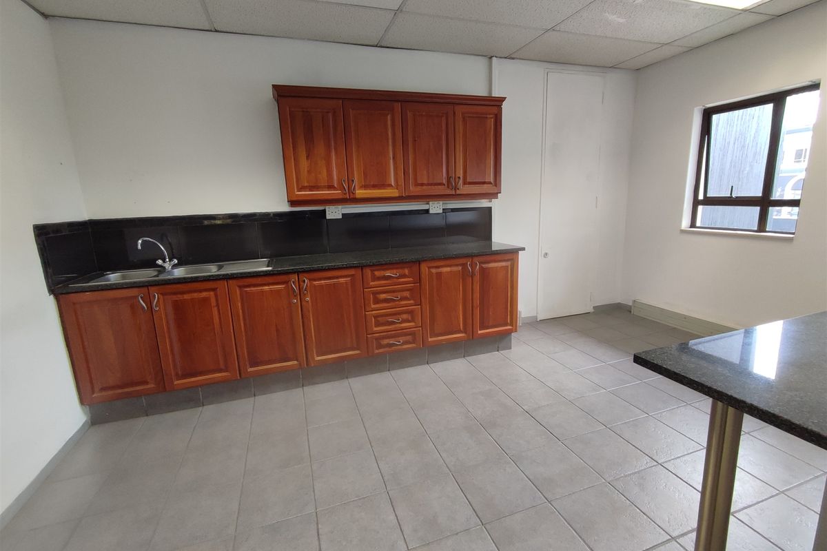 A functional office kitchenette featuring wooden cabinetry, granite countertops, a stainless steel sink, and neutral tiled flooring.