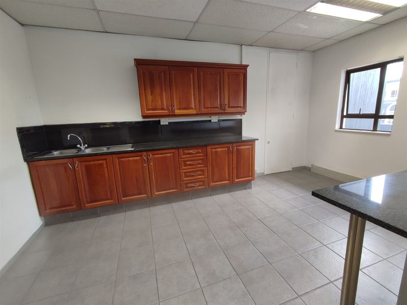 A functional office kitchenette featuring wooden cabinetry, granite countertops, a stainless steel sink, and neutral tiled flooring.