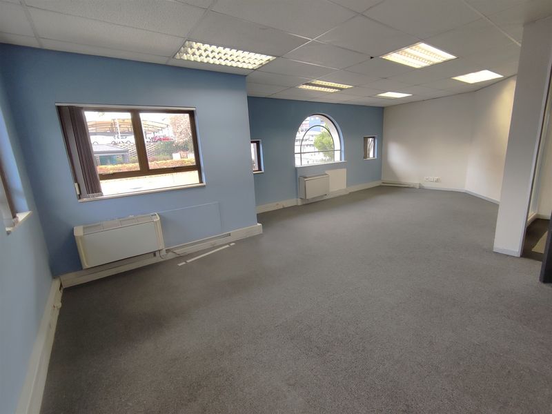 A spacious, empty office area featuring a suspended ceiling, recessed lighting, carpeted floors, and multiple windows providing natural light.