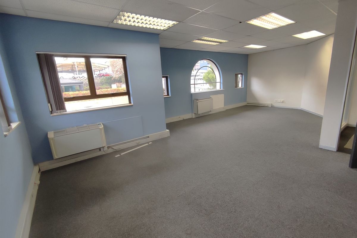 A spacious, empty office area featuring a suspended ceiling, recessed lighting, carpeted floors, and multiple windows providing natural light.