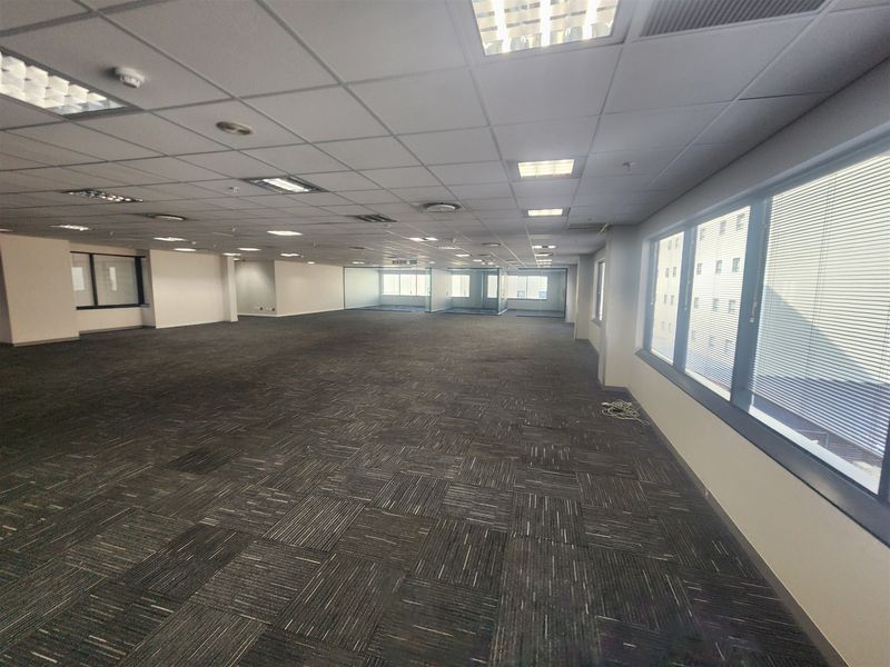 A spacious, open-plan office area featuring carpeted flooring, suspended ceilings with recessed lighting, and large windows with integrated blinds.