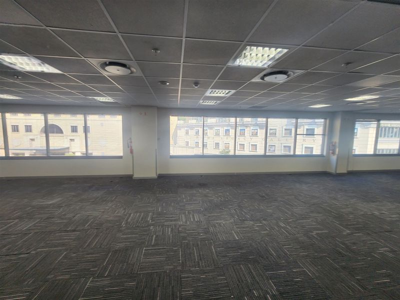 A spacious, empty open-plan office area featuring suspended ceilings, recessed lighting, and large windows providing ample natural light.