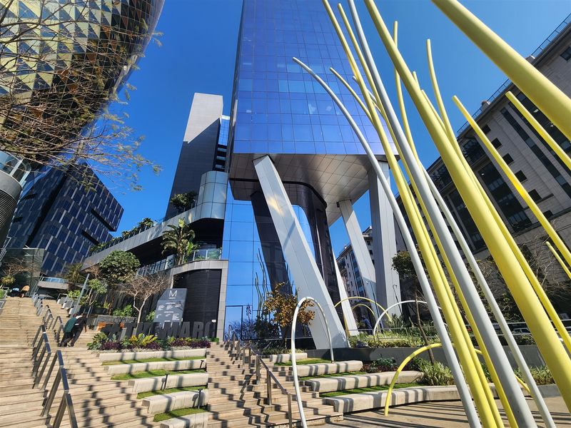 Modern commercial office building featuring a striking glass facade, elevated structural columns, and tiered landscaped pedestrian stairs in a contemporary urban plaza setting.