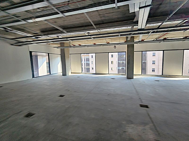 An expansive, open-plan office space in shell condition, featuring exposed ceiling infrastructure, concrete columns, and large windows providing ample natural light.