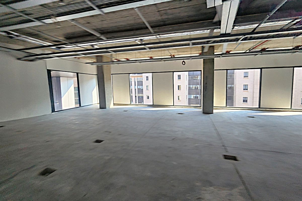 An expansive, open-plan office space in shell condition, featuring exposed ceiling infrastructure, concrete columns, and large windows providing ample natural light.