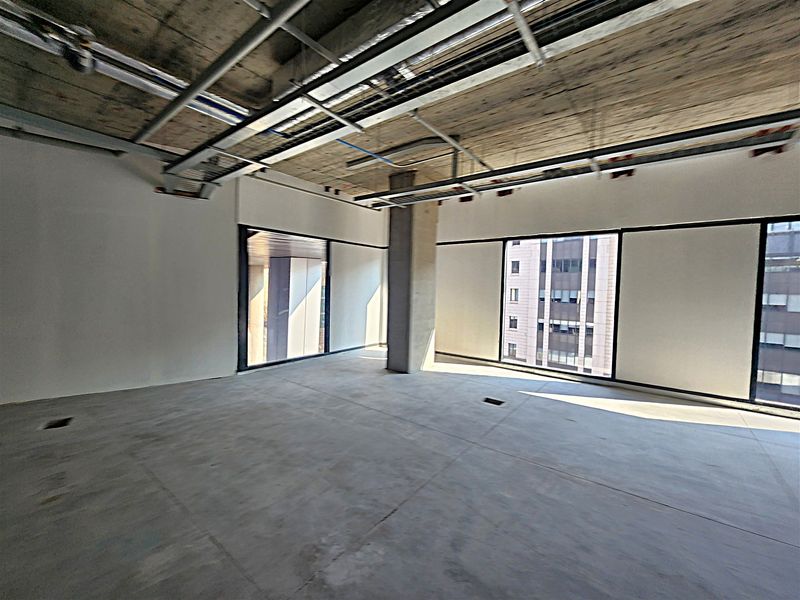 An open-plan office space in shell condition featuring exposed concrete ceilings, cable trays, and large windows providing ample natural light.
