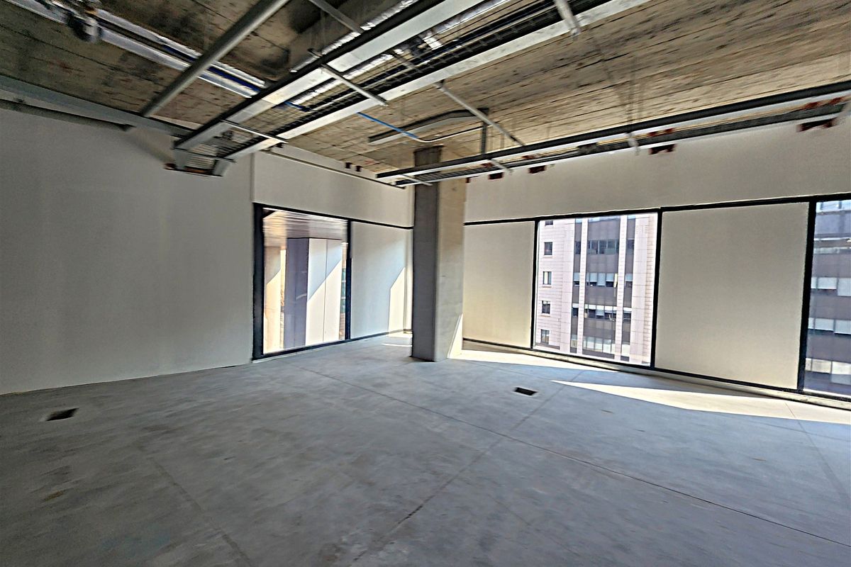 An open-plan office space in shell condition featuring exposed concrete ceilings, cable trays, and large windows providing ample natural light.