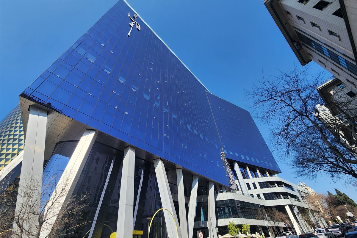 A modern high-rise office building featuring a striking blue glass curtain wall facade and prominent structural support columns.