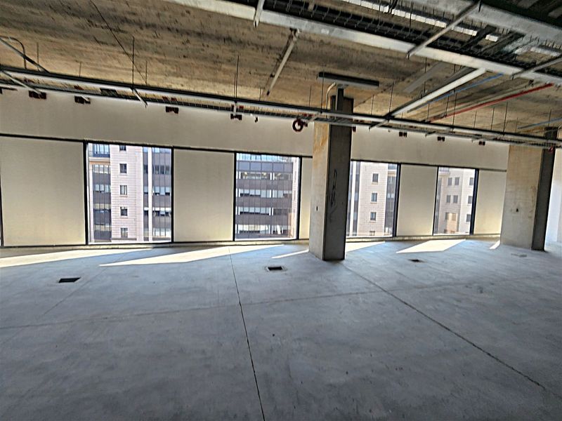An open-plan office space in shell condition featuring exposed concrete ceilings, structural columns, and large windows providing ample natural light.