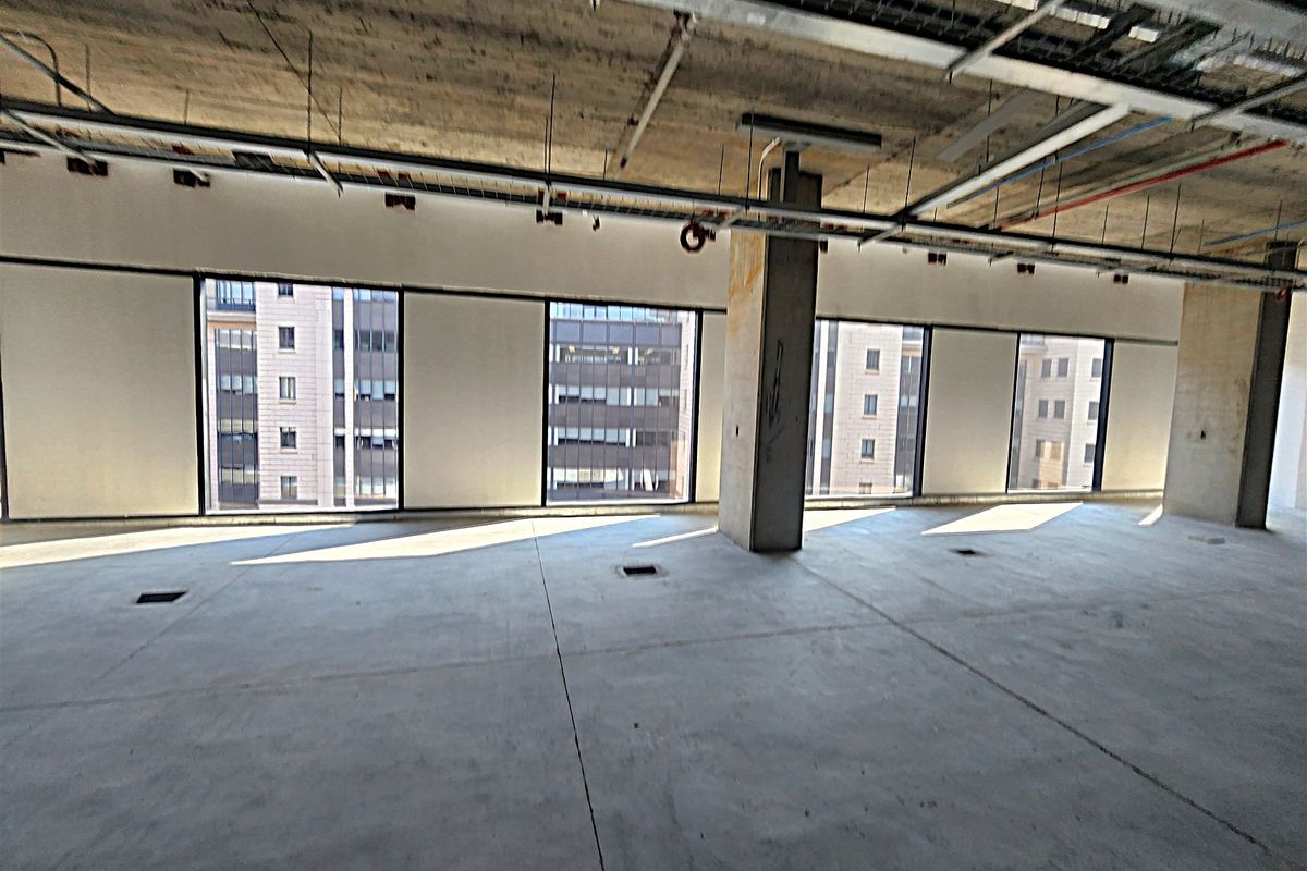 An open-plan office space in shell condition featuring exposed concrete ceilings, structural columns, and large windows providing ample natural light.
