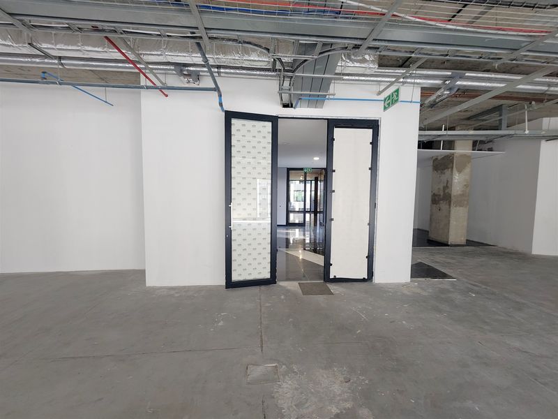 An unfinished open-plan office space featuring exposed ceiling infrastructure, concrete flooring, and a central double glass doorway.